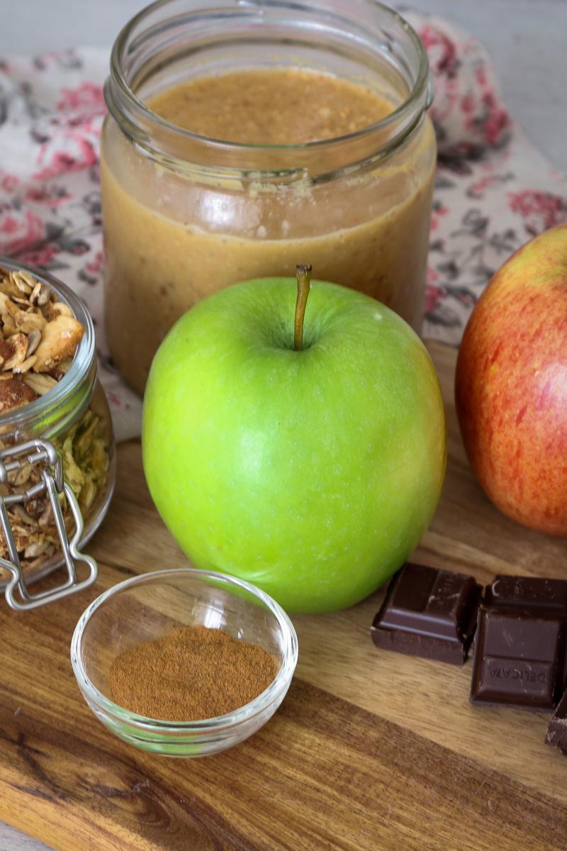 a green and red apple, a jar of almond butter, a cup fo granola, dairy free chocolate, and nut meg