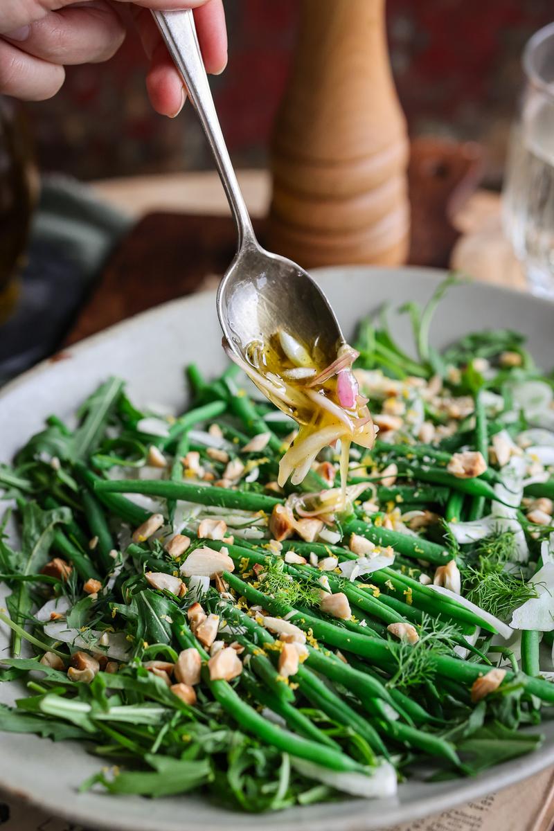 a plant-based person drizzling white wine vinagrette over a green bean salad recipe with shalots